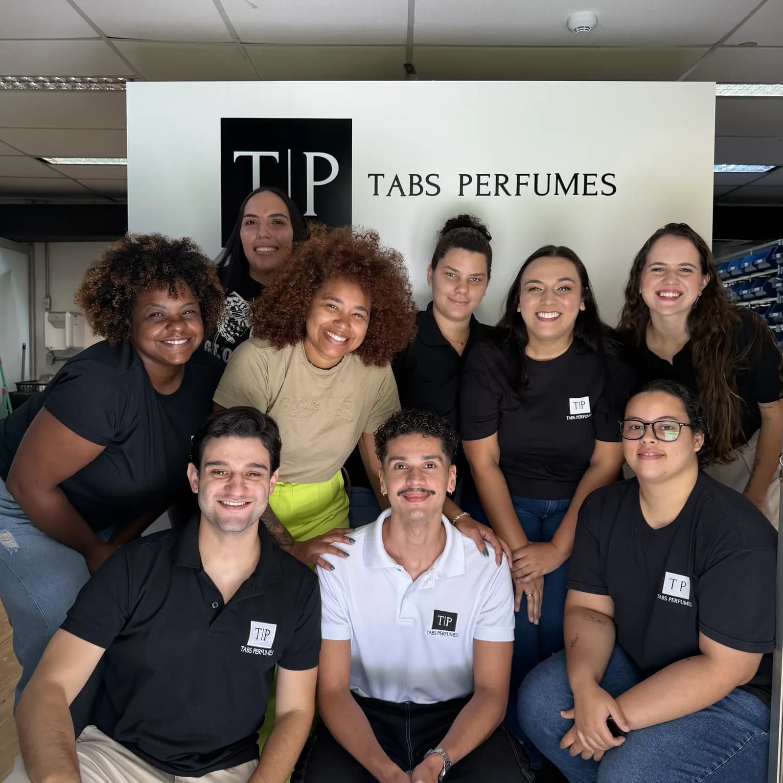 A equipe da Tabs reunida no escritório, diante do painel com o logo TP Tabs Perfumes