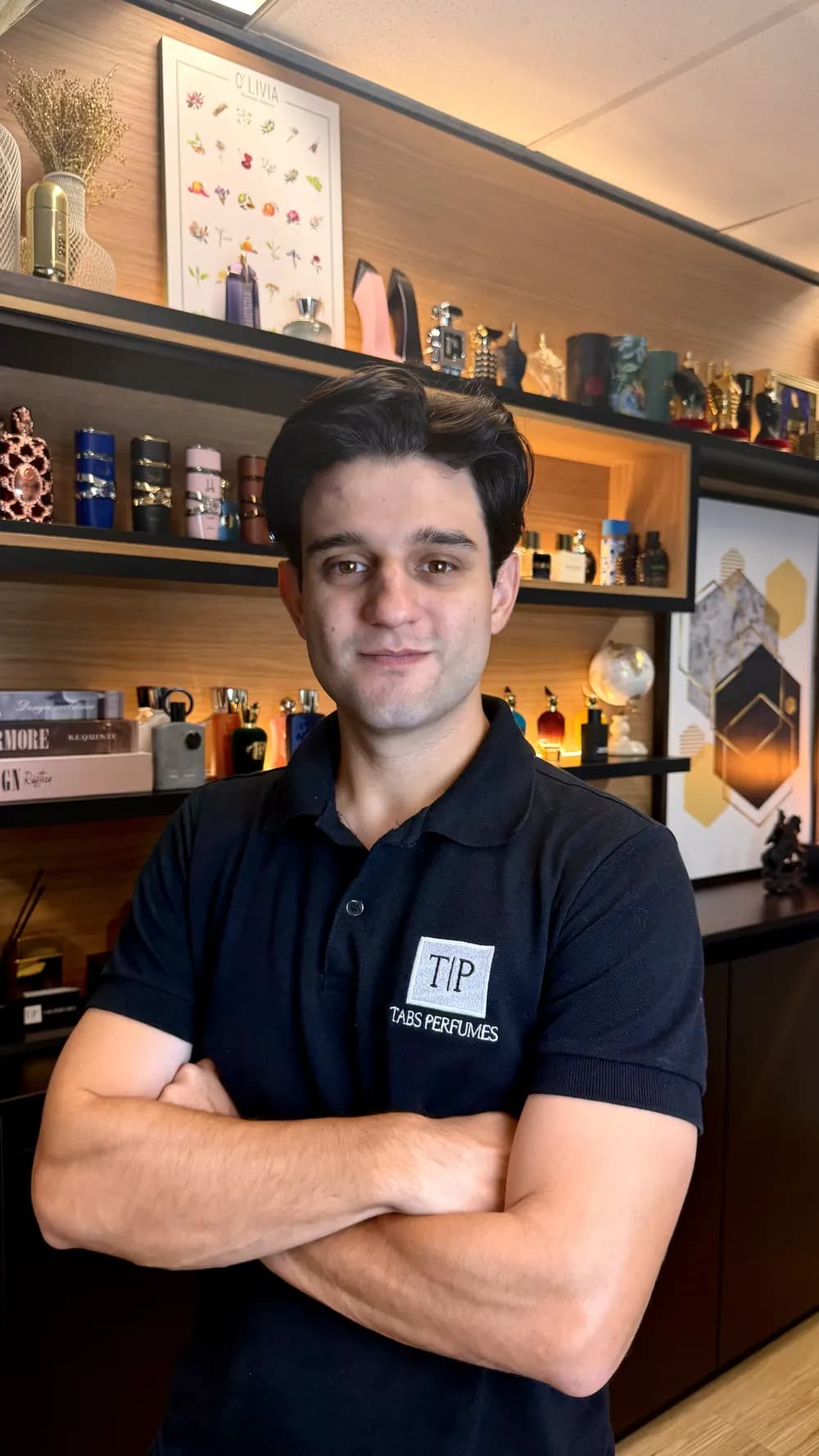 Gabriel Tabchoury, fundador da Tabs, de braços cruzados com a camisa da Tabs, diante da estante de perfumes